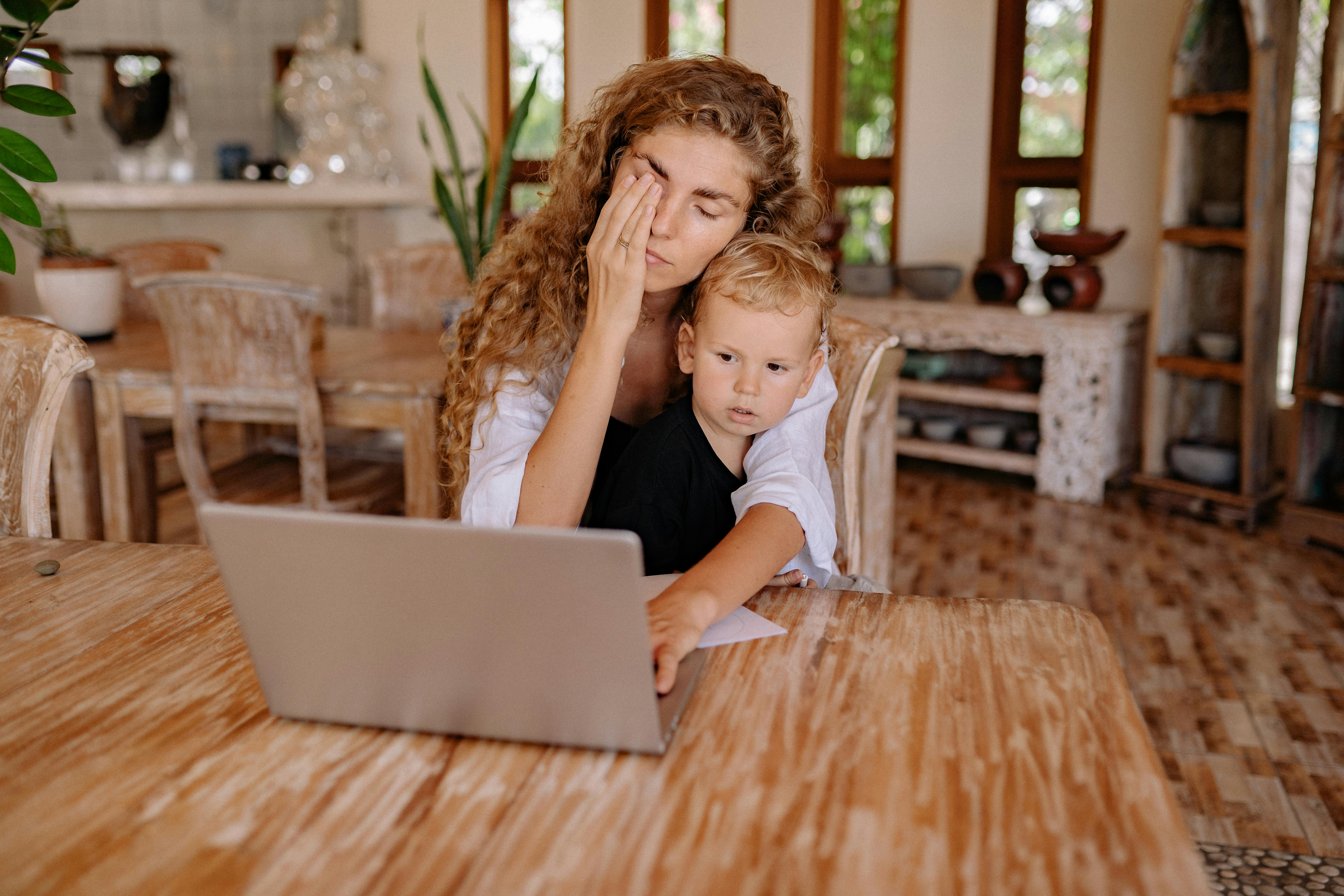 Tired mom with toddler on her lap rubs sleep from her eyes in front of a laptop