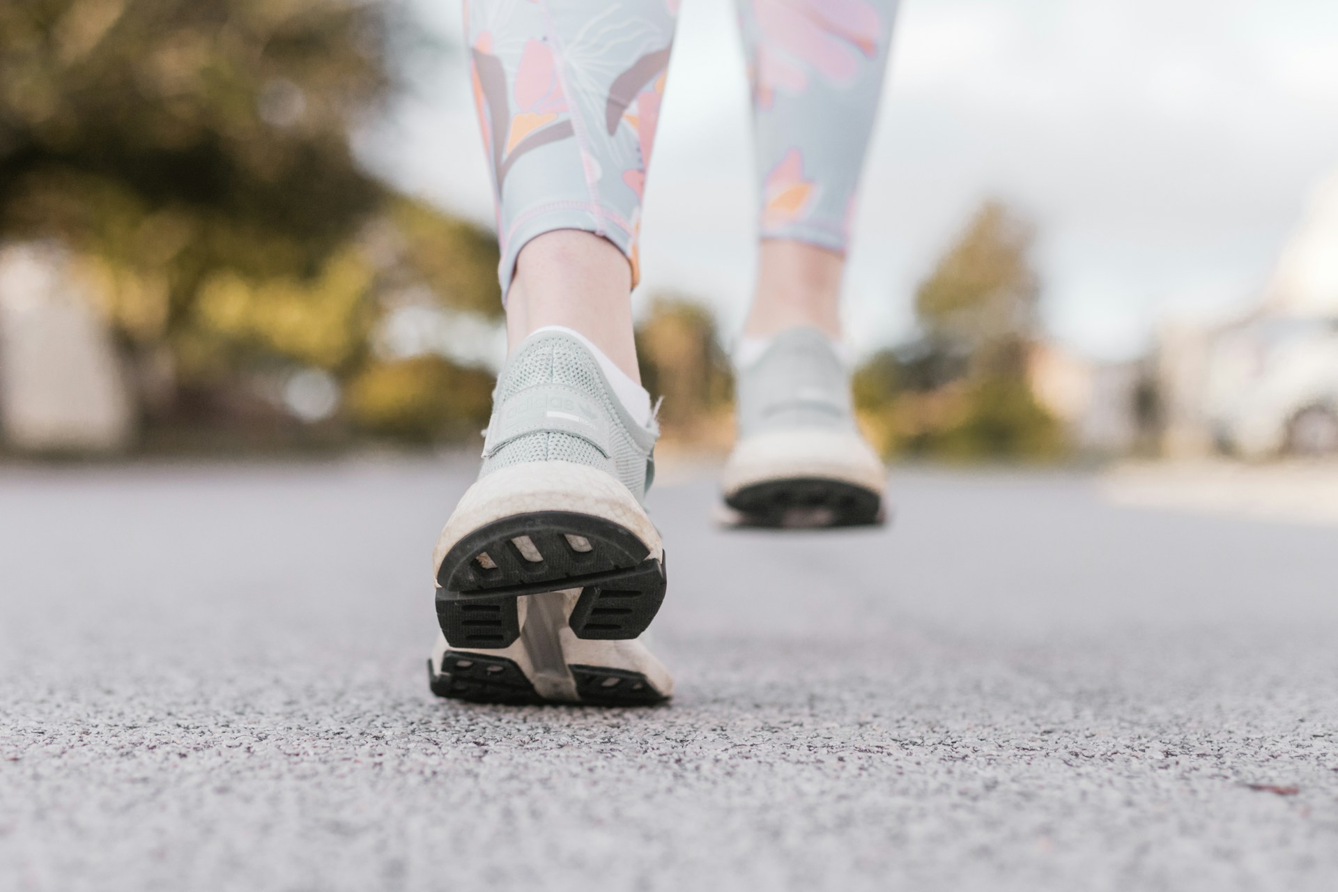 Low-angle close-up of a person walking on a paved road, focusing on the soles of athletic sneakers mid-step, with blurred trees and suburban surroundings in the background under soft natural daylight.