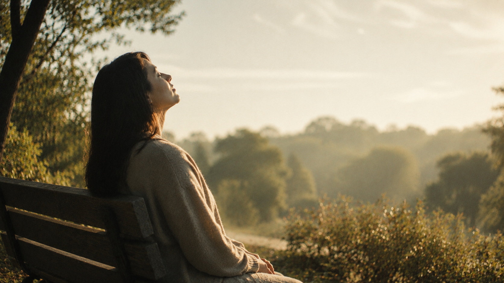 A person sitting quietly on a wooden bench in soft early morning sunlight, eyes closed and face turned gently toward the light. Warm golden tones, blurred trees, and subtle film grain create a peaceful atmosphere of stillness, reflection, and quiet renewal.