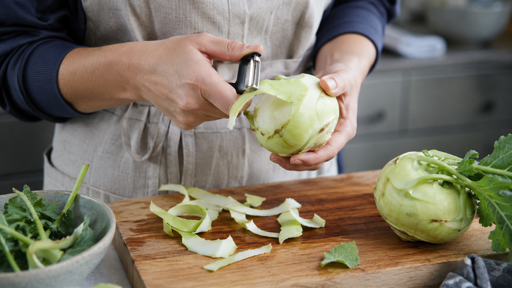A person uses a vegetable peeler to remove the outer skin from a fresh kohlrabi bulb over a wooden cutting board.