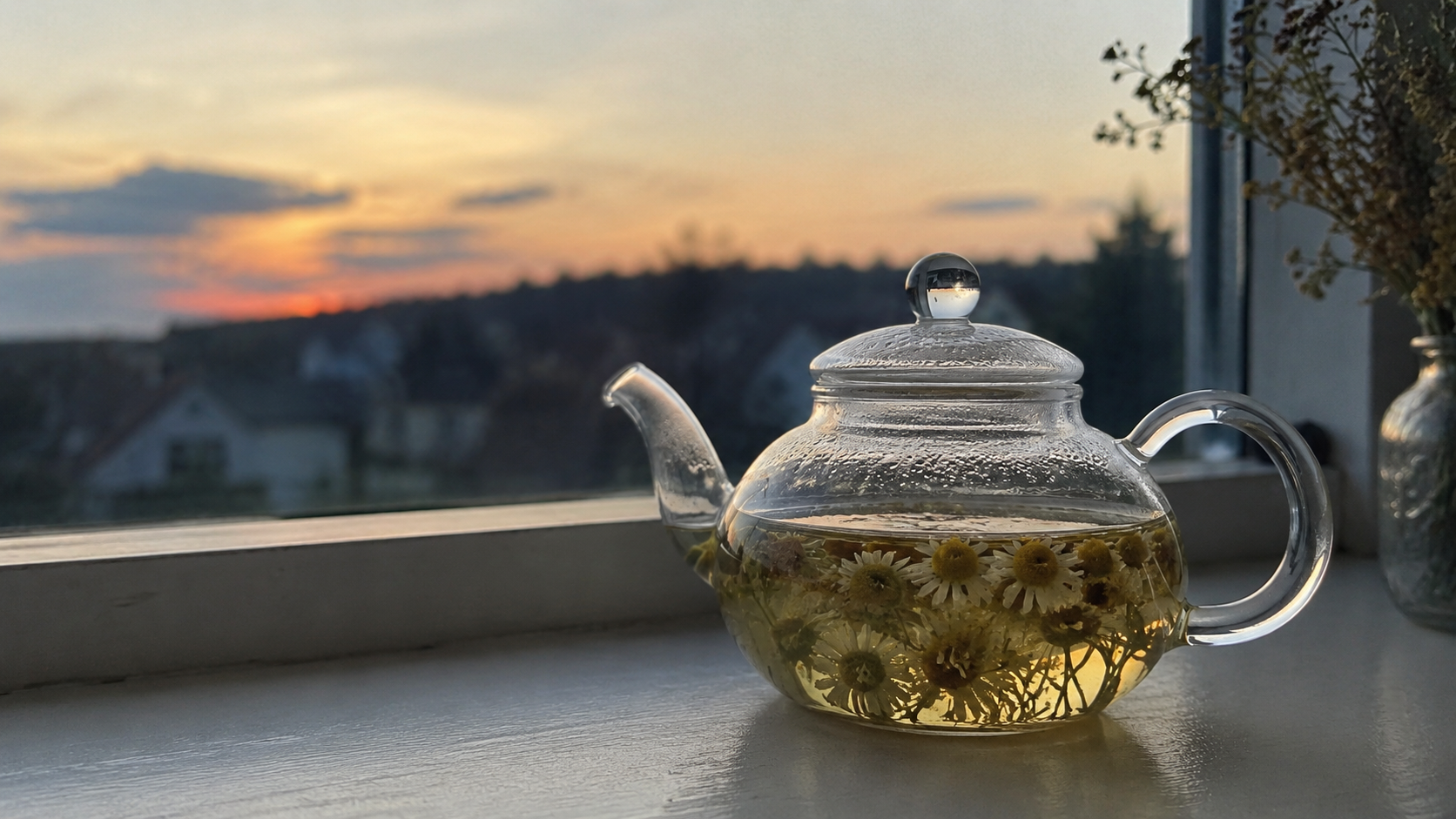 A clear glass teapot filled with golden chamomile tea and floating flowers rests on a windowsill against a softly glowing sunset.