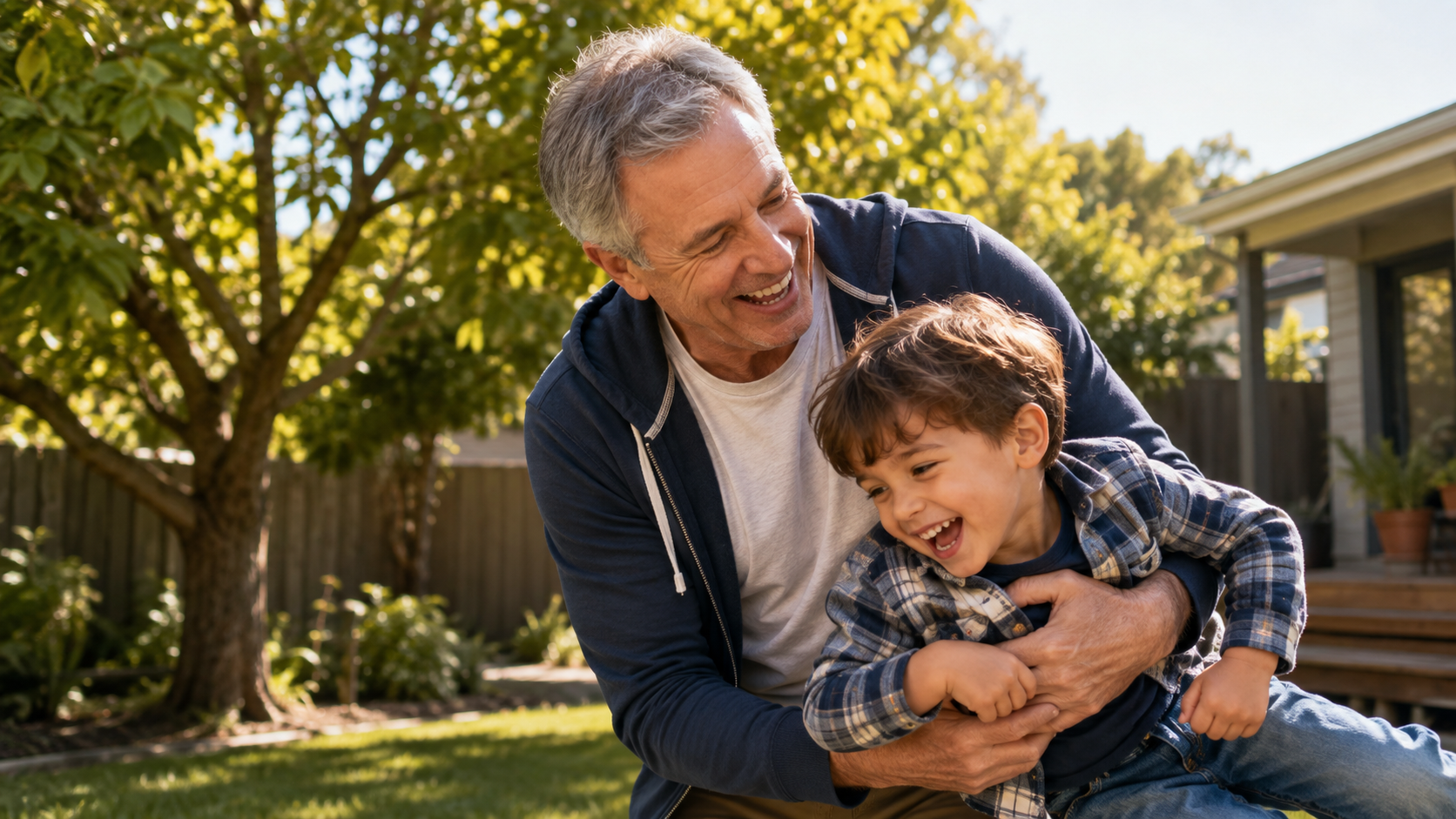 A smiling older man plays with his young grandson in a sunny backyard, sharing a joyful and energetic moment together.