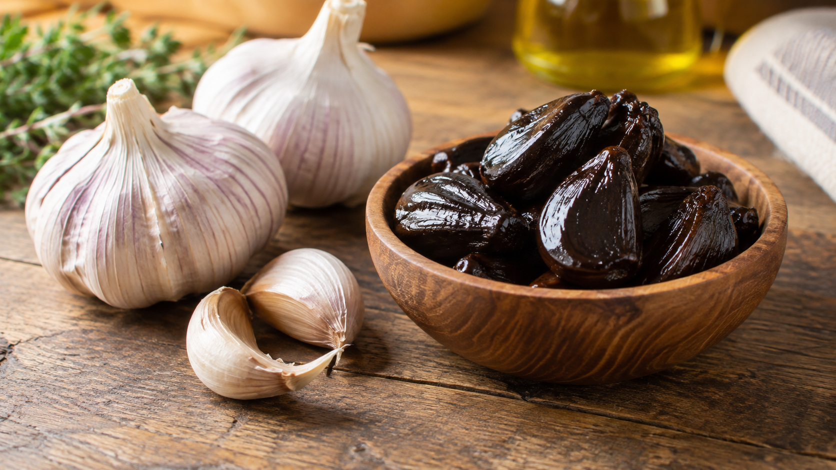 A wooden bowl filled with glossy black garlic cloves sits beside whole bulbs and loose cloves of fresh raw garlic on a rustic wooden table in soft natural light.